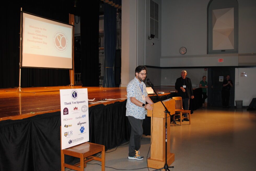 A speaker presents at a podium in a conference room with a projection screen and an informational sign.