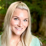 Smiling blonde woman outdoors with greenery in background.